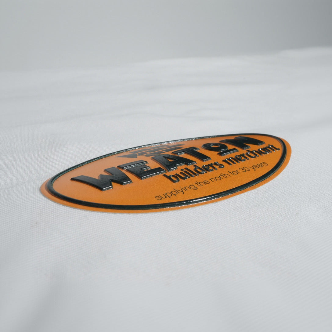 An oval orange and black tpu patch with the text 'WEATON builders merchant' raised and 'supplying the north for 30 years' on a white textured fabric background.