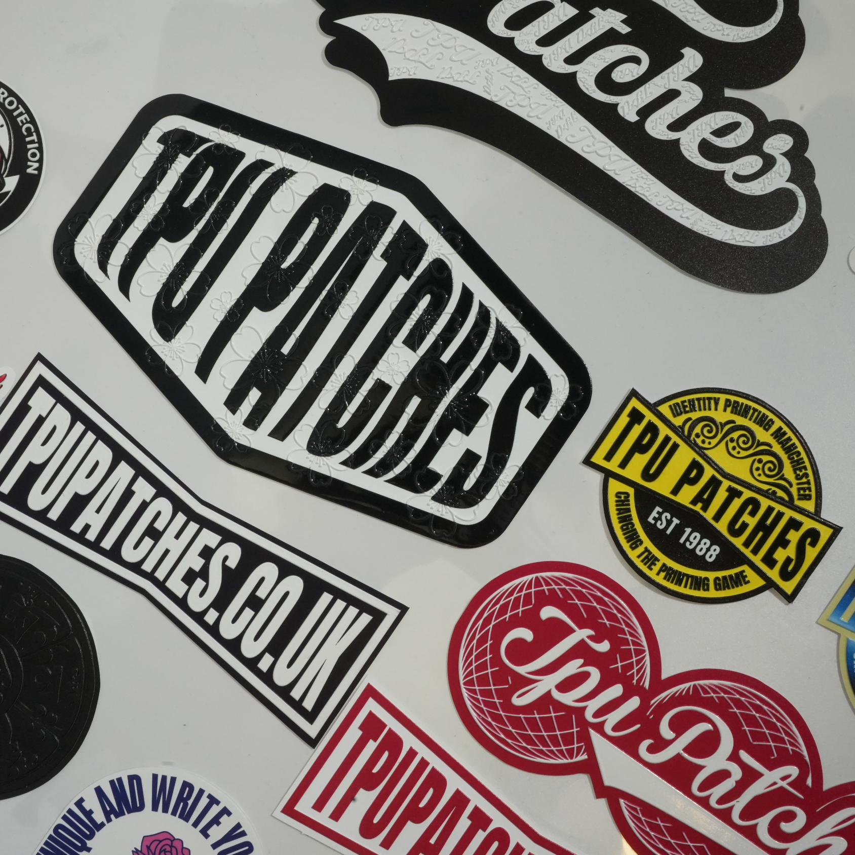 A close-up of multiple TPU Patches brand transfers arranged on a white surface, including a large black-and-white central transfer, a red-and-white heart transfer, a yellow circular badge transfer, and other colourful graphic transfer.