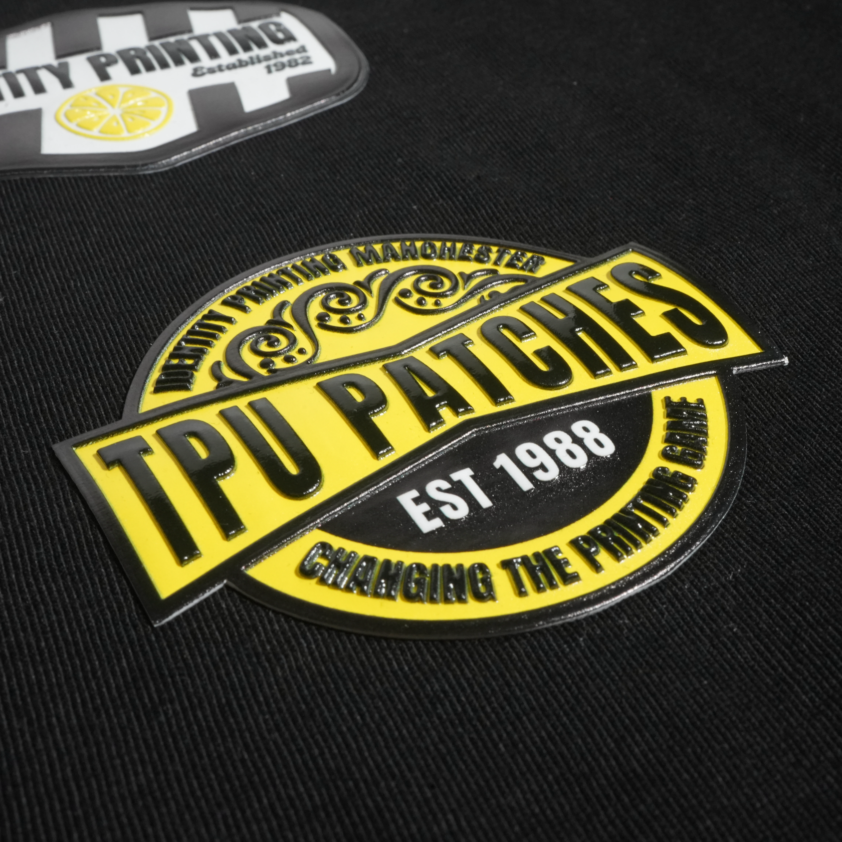 A close-up of a black fabric surface featuring two patches. The main patch is a large yellow and black oval with the text 'TPU PATCHES' and 'EST 1988'.